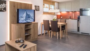 Eine moderne Küche mit Einbauofen und Mikrowelle, ein Essbereich mit Holztisch und Stühlen sowie ein an einer Holzkommode befestigter Fernseher.