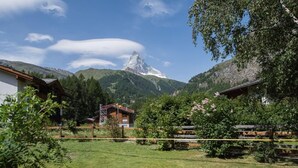 Ländliche Szenerie mit Berg im Hintergrund, einem Holzhaus und einem umzäunten Garten.