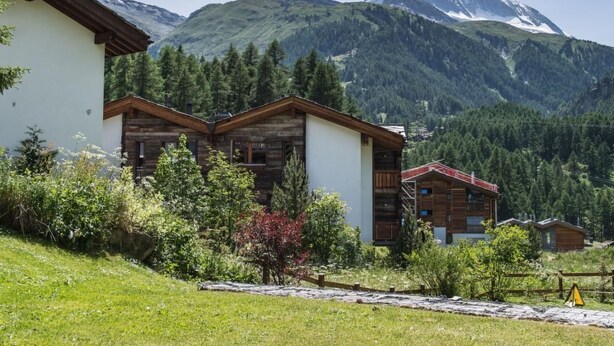 A chalet-style building with a wooden exterior and white walls, surrounded by lush greenery and a mountain backdrop. - Top Ferienwohnung Monte Rosa (SkiIn/SkiOut) by Interhome (Zermatt)