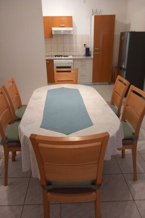 A dining area with a table set for six, surrounded by wooden chairs with green cushions. A kitchen with a stove, oven, and cabinets is visible in the background. - "Apartment Mate 1" am Strand by Interhome (Brač/Bol)
