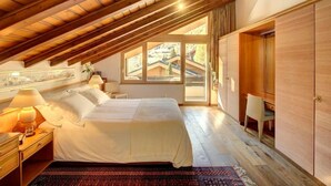 Een slaapkamer met schuine plafonds, een bed, een nachtkastje, een lamp, een raam met uitzicht op een gebouw en een kledingkast.