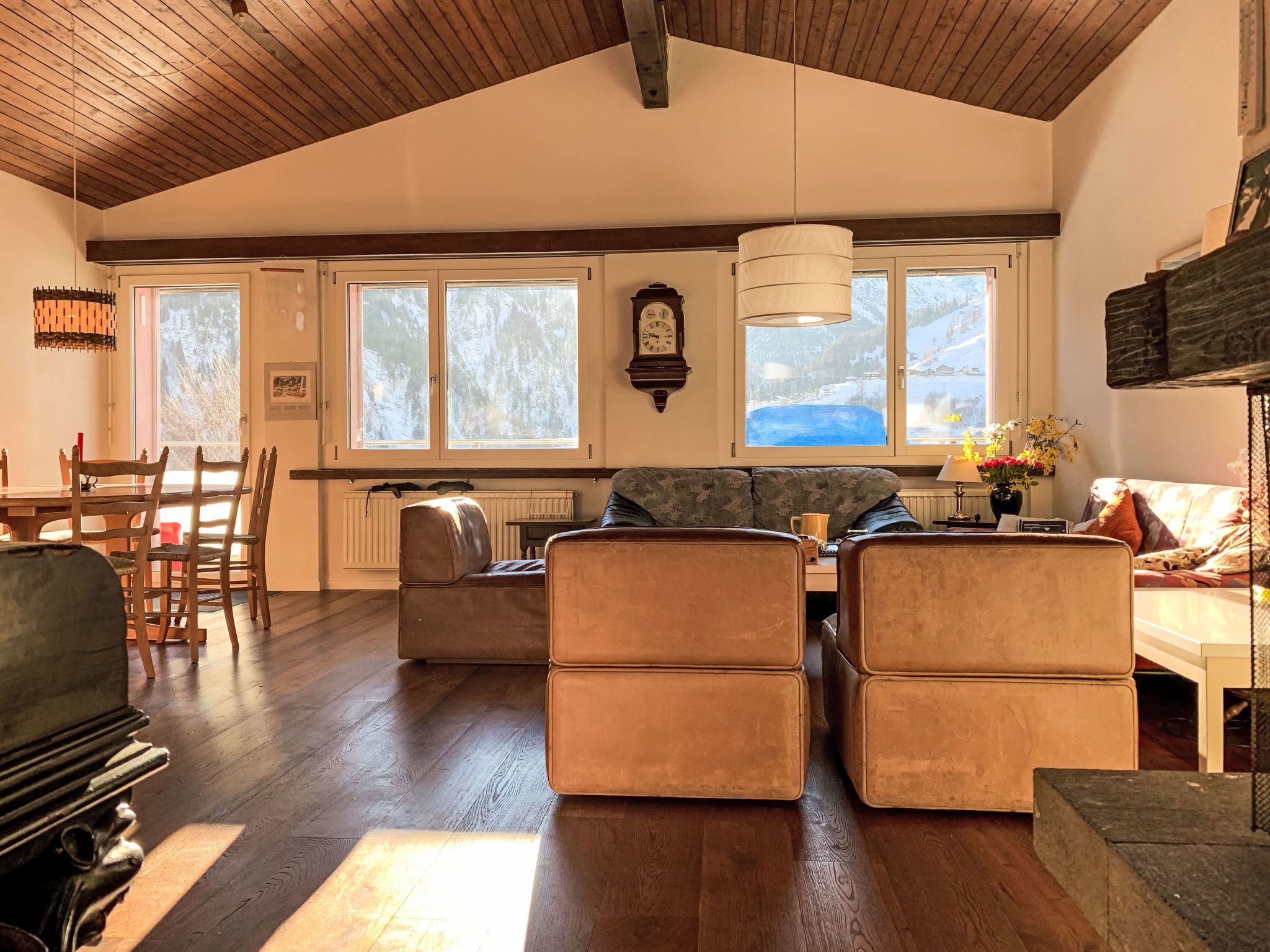 Un vaste espace de vie doté d’un haut plafond en bois, d’une cheminée, d’une table à manger avec des chaises et de grandes fenêtres offrant une vue sur les arbres.