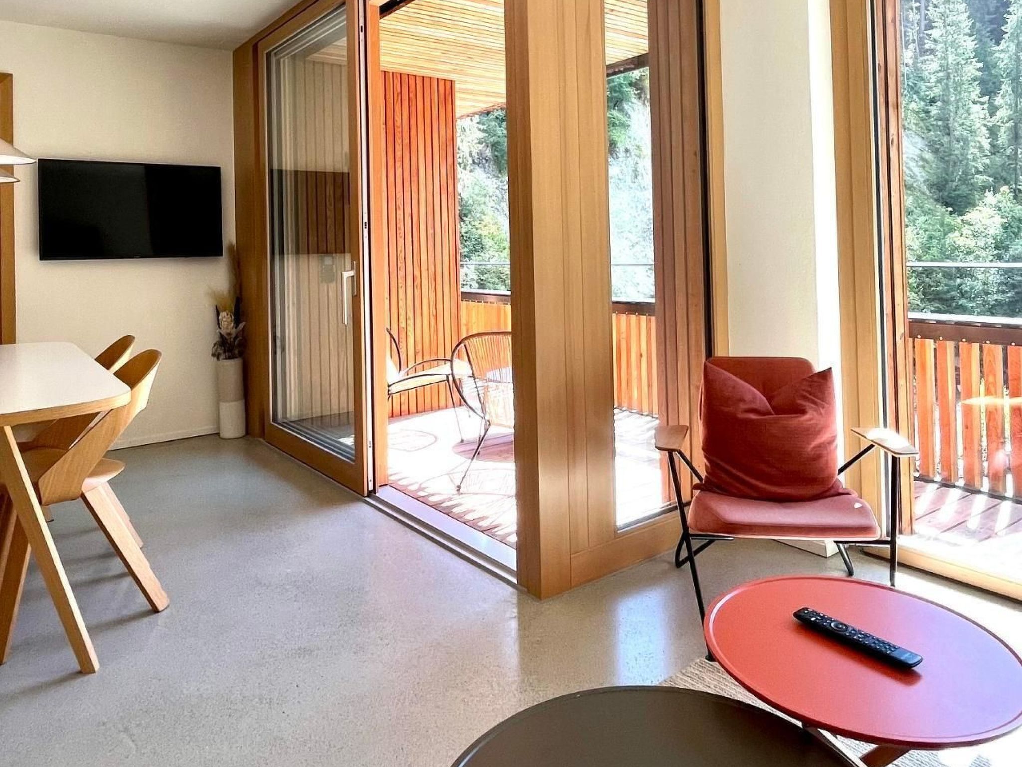 Une chambre moderne dotée d’une porte-fenêtre coulissante donnant sur un espace extérieur, d’un téléviseur à écran plat fixé au mur, d’une table à manger avec des chaises, et d’un fauteuil rouge avec une télécommande posée sur une petite table.