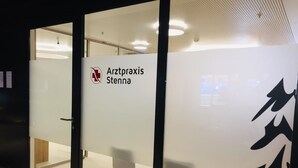 Et moderne interiør med en glasdør med teksten "Arztpraxis Stenna". Indenfor er der to hvide stole og et trægulv.