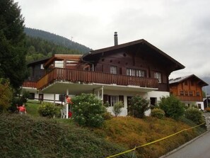 Un chalet à deux étages doté d’un balcon, entouré de verdure et adossé à une montagne boisée.
