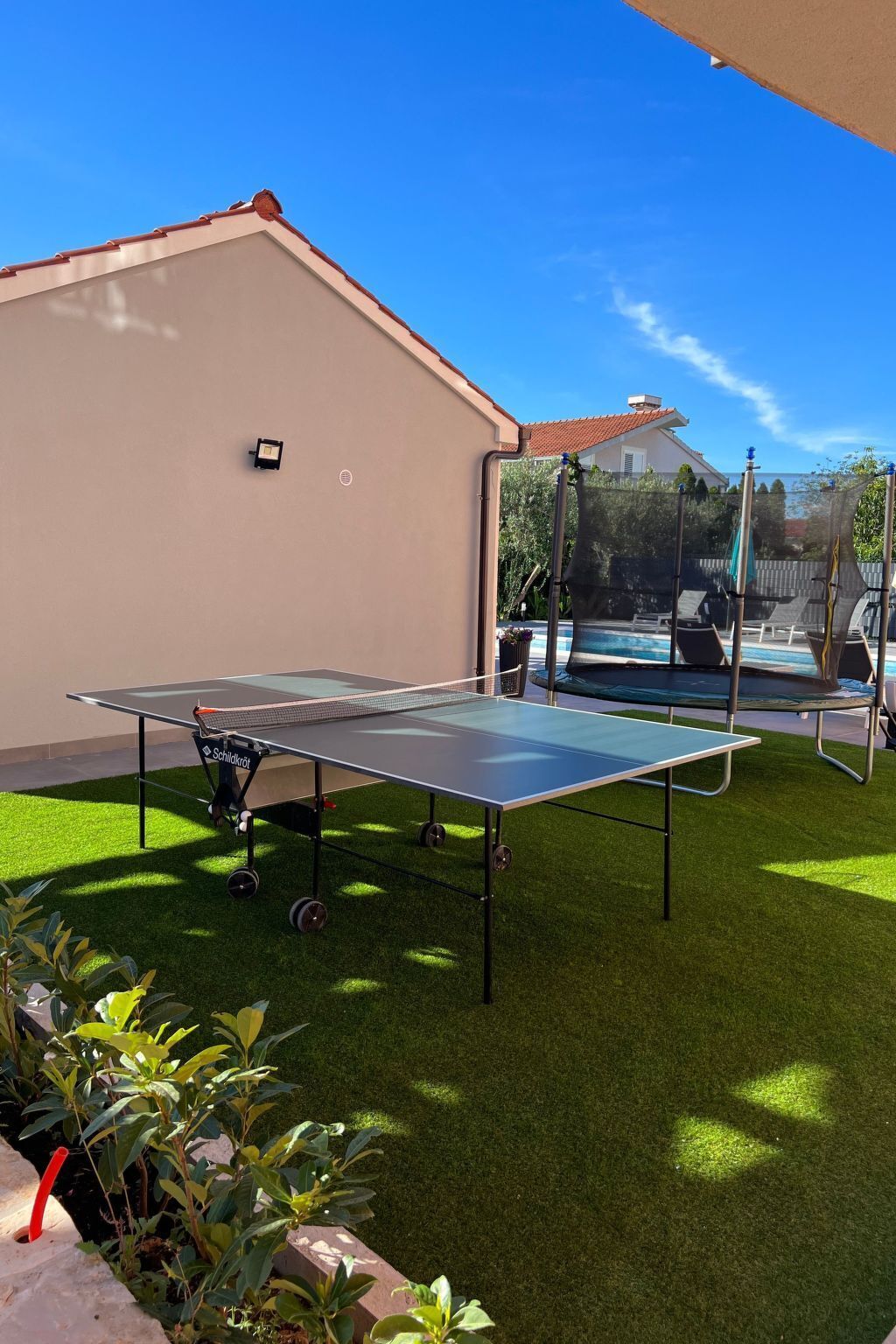 An outdoor ping pong table set up on artificial grass with a pool in the background.