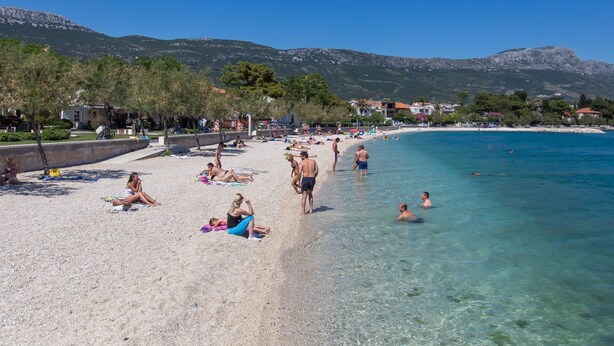 Une plage avec une eau turquoise et limpide, des gens qui se prélassent au soleil, et des montagnes en arrière-plan.