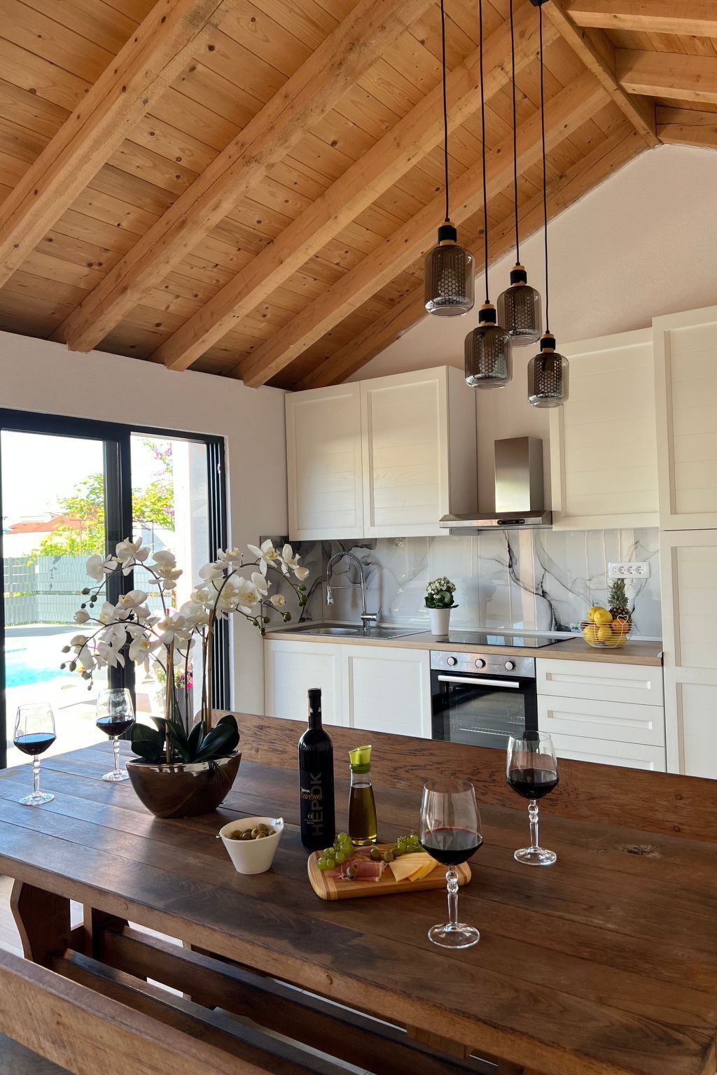 A modern kitchen with wooden beams, white cabinets, and a wooden dining table set with wine glasses, a bottle, and a fruit platter.