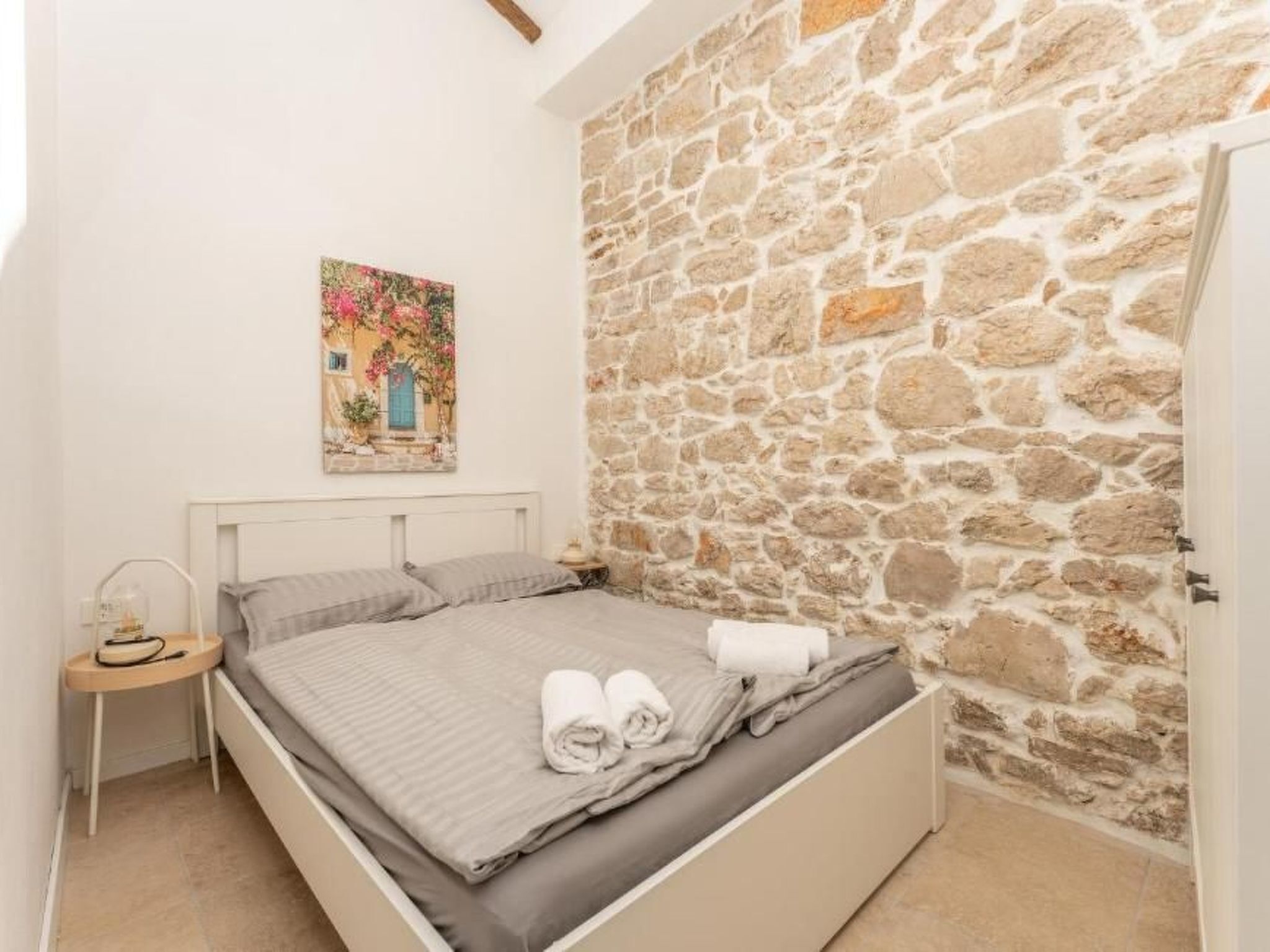 Une chambre avec un mur en pierre, un lit avec une literie grise, une table de chevet avec une lampe et un tableau accroché au mur.