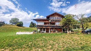 A two-story wooden house with a balcony, surrounded by a grassy field and trees. - Ferienwohnung mit Bergpanorama by Interhome (Zweisimmen)