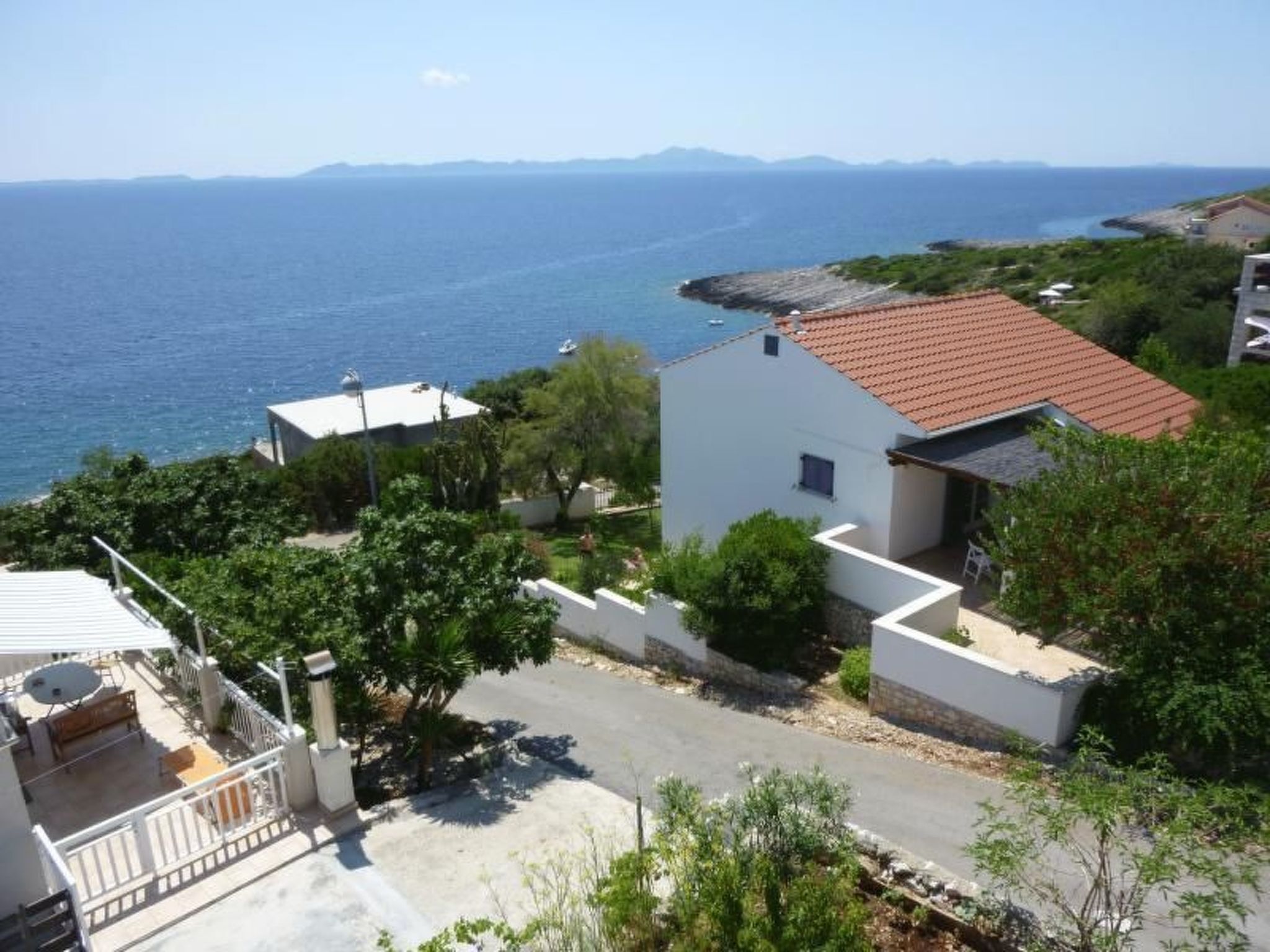 A coastal residential area with a white house featuring a red-tiled roof, surrounded by greenery and a clear view of the sea.