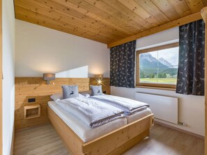 A bedroom with a wooden bed, two bedside tables, a nightstand, a window with curtains, and a view of mountains. - Appartement "Hochgefühl" by Interhome (Leutasch)