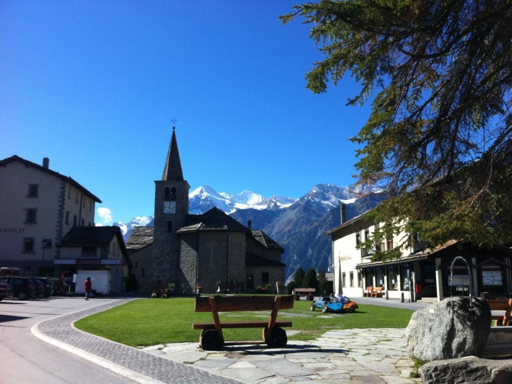 Eine Steinsitzbank auf einem Stadtplatz, im Hintergrund eine Kirche und Berge.