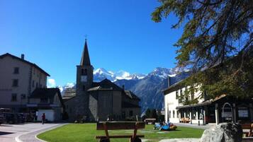 Eine Steinsitzbank auf einem Stadtplatz, im Hintergrund eine Kirche und Berge.