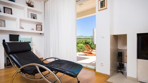 Una habitación con una butaca reclinable negra, un televisor, una estantería con fotografías enmarcadas y vistas al exterior con vegetación.