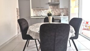 Et moderne køkken med lysegrå skabe, en rustfri emhætte og et flisebeklædt bagpanel. Der er et spisebord med hvid dug og to stole.
