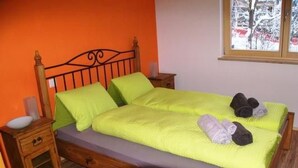 A bed with a wooden frame, a green bedspread, and pillows. There is a nightstand with a lamp and a small drawer.