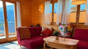 A cozy cabin interior with a red sofa, a wooden table with a plant, and a view of mountains through large windows. - Chalet Poiema by Interhome (Bellwald)