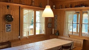 A wooden cabin with a dining table, chairs, lace curtains, and a hanging light fixture. - Chalet Glücksfall by Interhome (Bellwald)