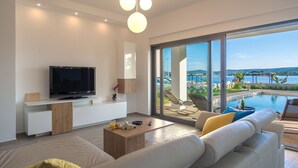 Ett modernt vardagsrum med en stor platt-TV, en soffa, ett soffbord och utsikt över en pool och havet.
