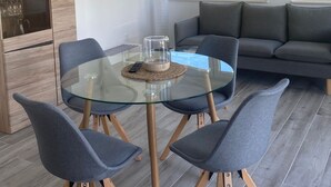 Un comedor moderno con una mesa de vidrio, cuatro sillas grises, un sofá gris y una lámpara colgante.
