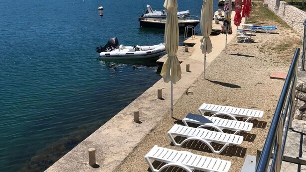 A waterfront area with lounge chairs and umbrellas, a dock with boats, and a forested hillside in the background. - Ferienwohnung by Interhome (Novigrad (Zadar))