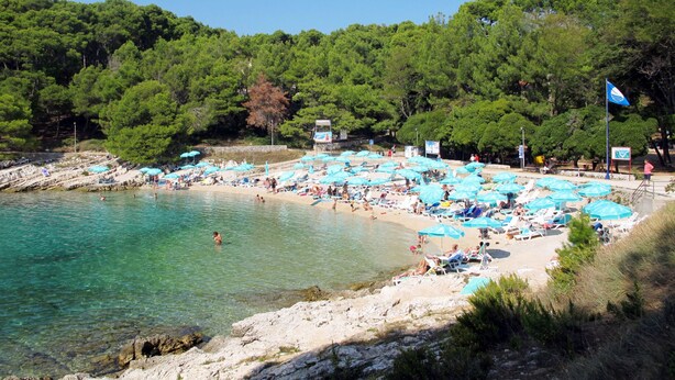 Ein Strand mit klarem türkisblauem Wasser, vielen blauen Sonnenschirmen und Menschen, die sich sonnen.