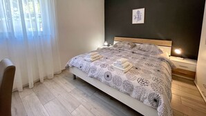 Ein Schlafzimmer mit Bett, Nachttisch mit Lampe, einem Sessel und einem Fenster mit durchsichtigen Vorhängen.