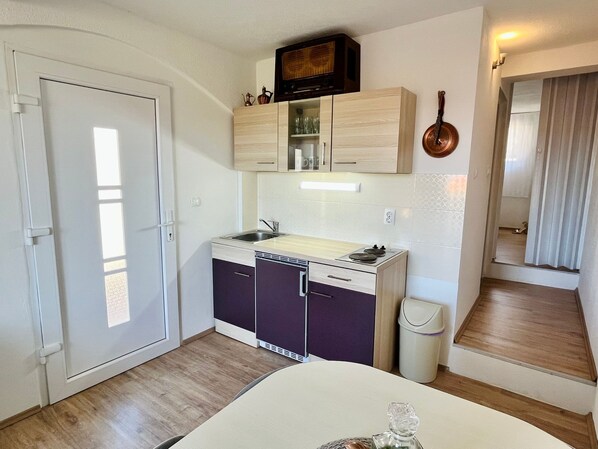 Cocina compacta con lavamanos, estufa y alacenas, un basurero, y una puerta que da a otra habitación.