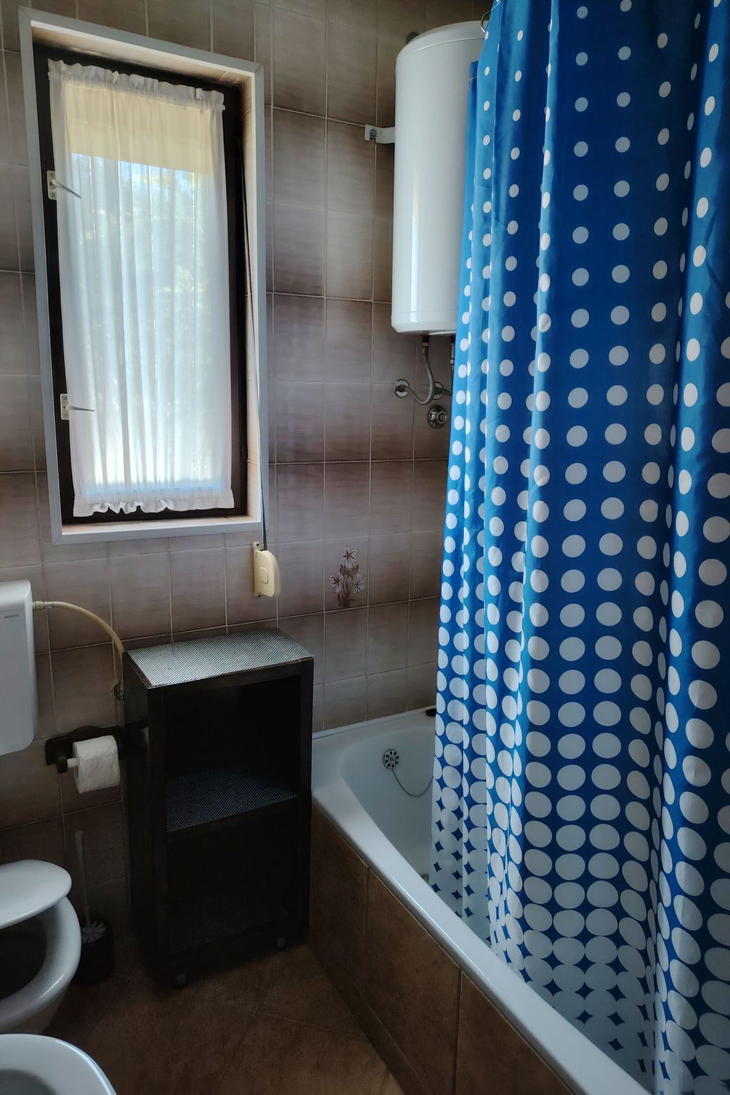 A bathroom with a toilet, a small sink, a window with a white curtain, and a bathtub with a blue polka dot shower curtain.