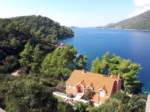 Uma casa costeira com telhado cerâmico, rodeada por vegetação densa e vista para o mar.