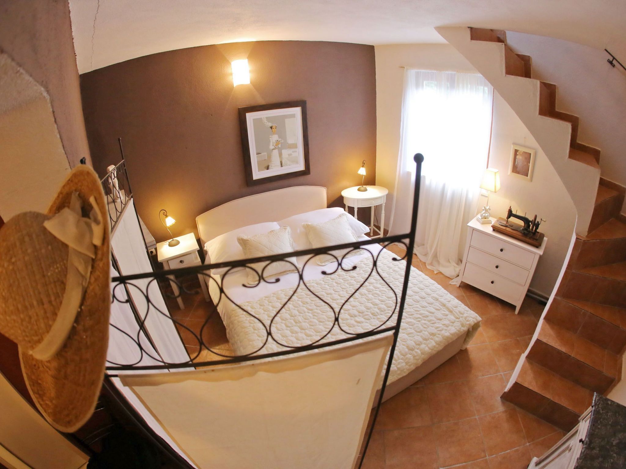 A bedroom with a curved ceiling, a bed with a white headboard, a nightstand, a small table with a lamp, a sewing machine on a dresser, and a staircase.