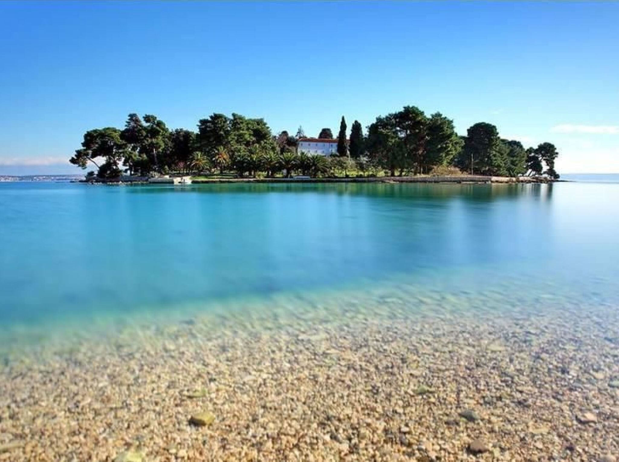 A clear view of a pebble beach leading to a small island with trees and a building.