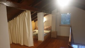 A bedroom with a wooden ceiling, a bed with floral bedding, a nightstand, a window with blinds, and a staircase. - Am Fluss gelegenes Haus mit Galerieschlafplätzen by Interhome (Nin)