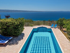 Piscina con vista al mar, rodeada de vegetación y sillones de descanso.