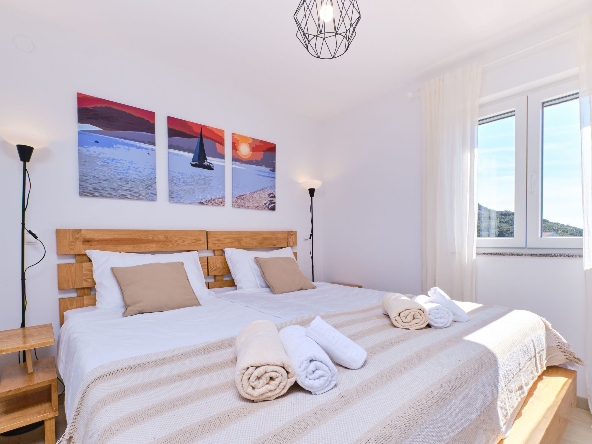 A neatly made bed with rolled towels, a wooden headboard, and three sunset paintings on the wall.