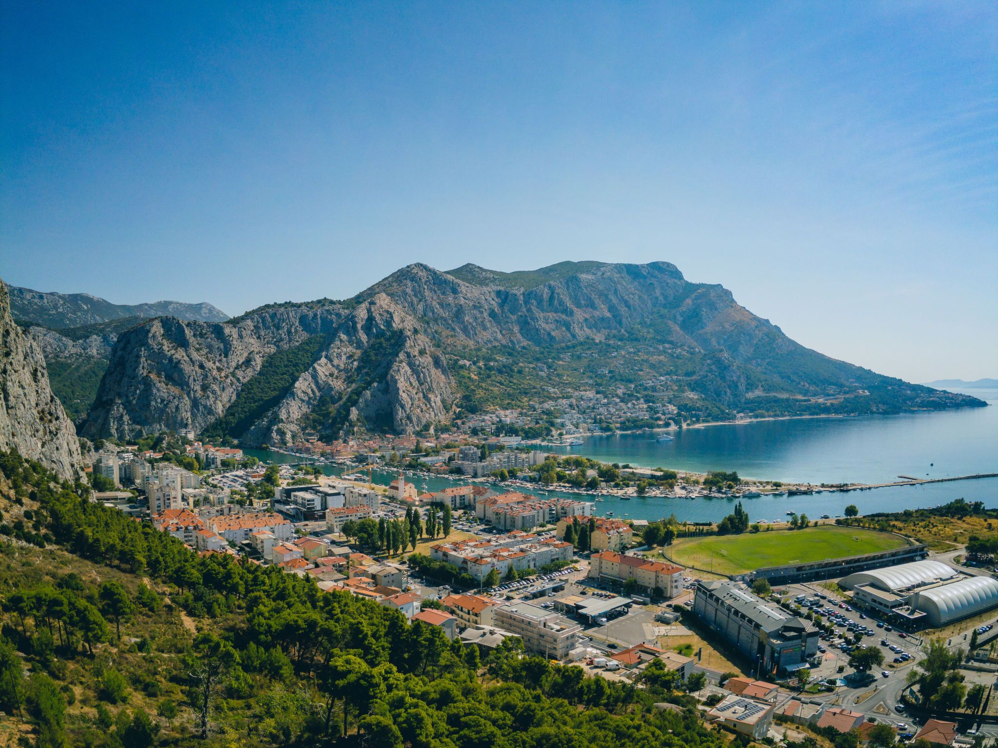 A coastal town with mountainous terrain in the background, a marina, and a large stadium.