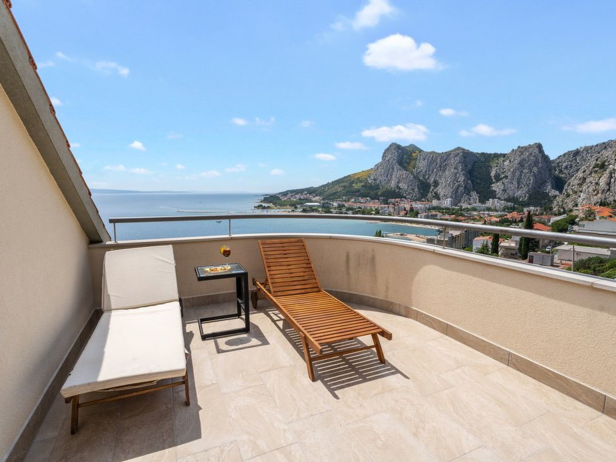 A balcony with a wooden lounge chair and a small table, overlooking a coastal town and mountains.
