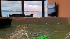 Habitación con un ventanal grande con vistas a un cuerpo de agua y montañas, una bañera con luces verdes y un televisor en la pared.