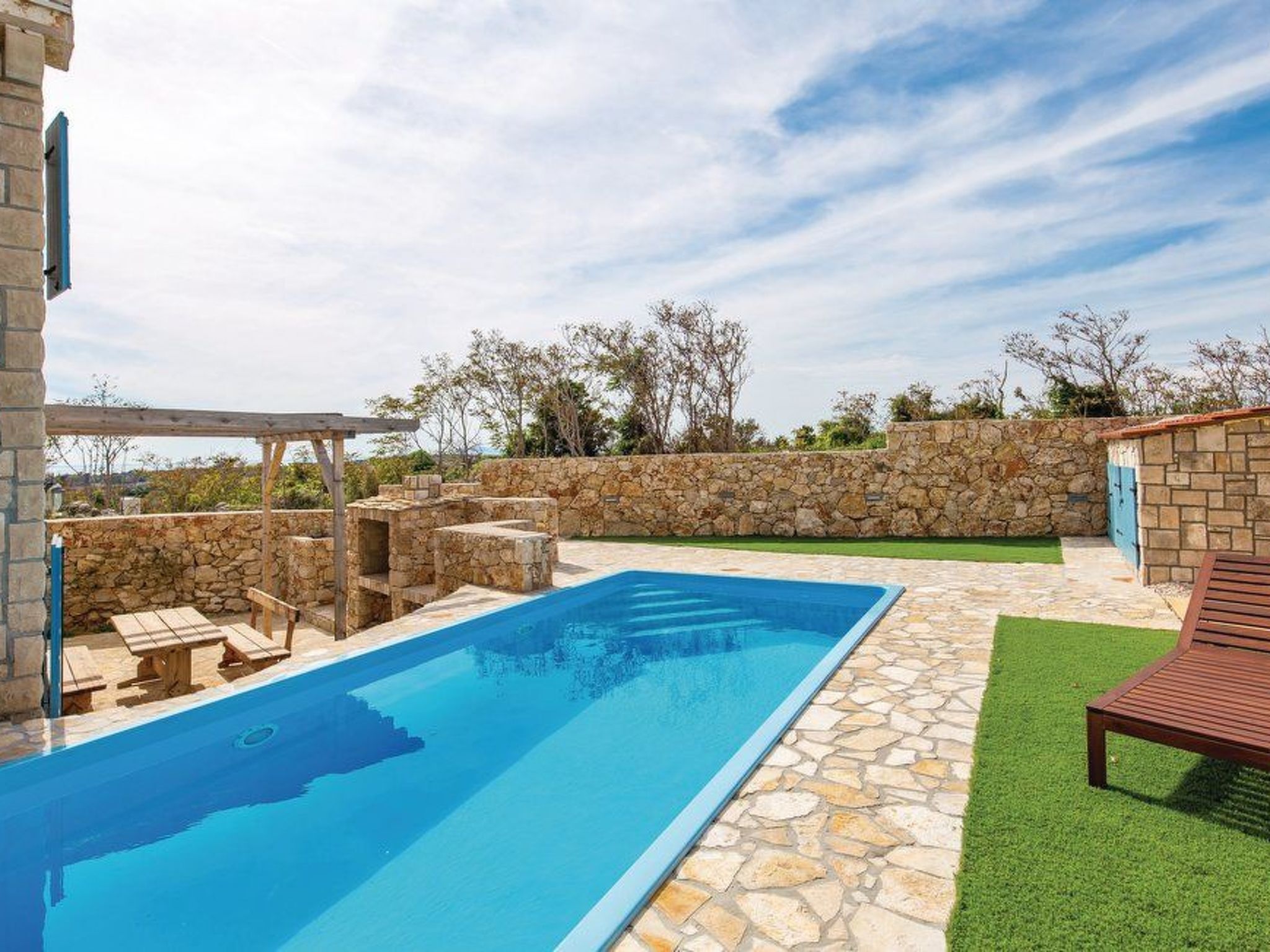 A swimming pool with stone tiles, wooden deck seating, and a pergola.