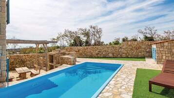 A swimming pool with stone tiles, wooden deck seating, and a pergola.