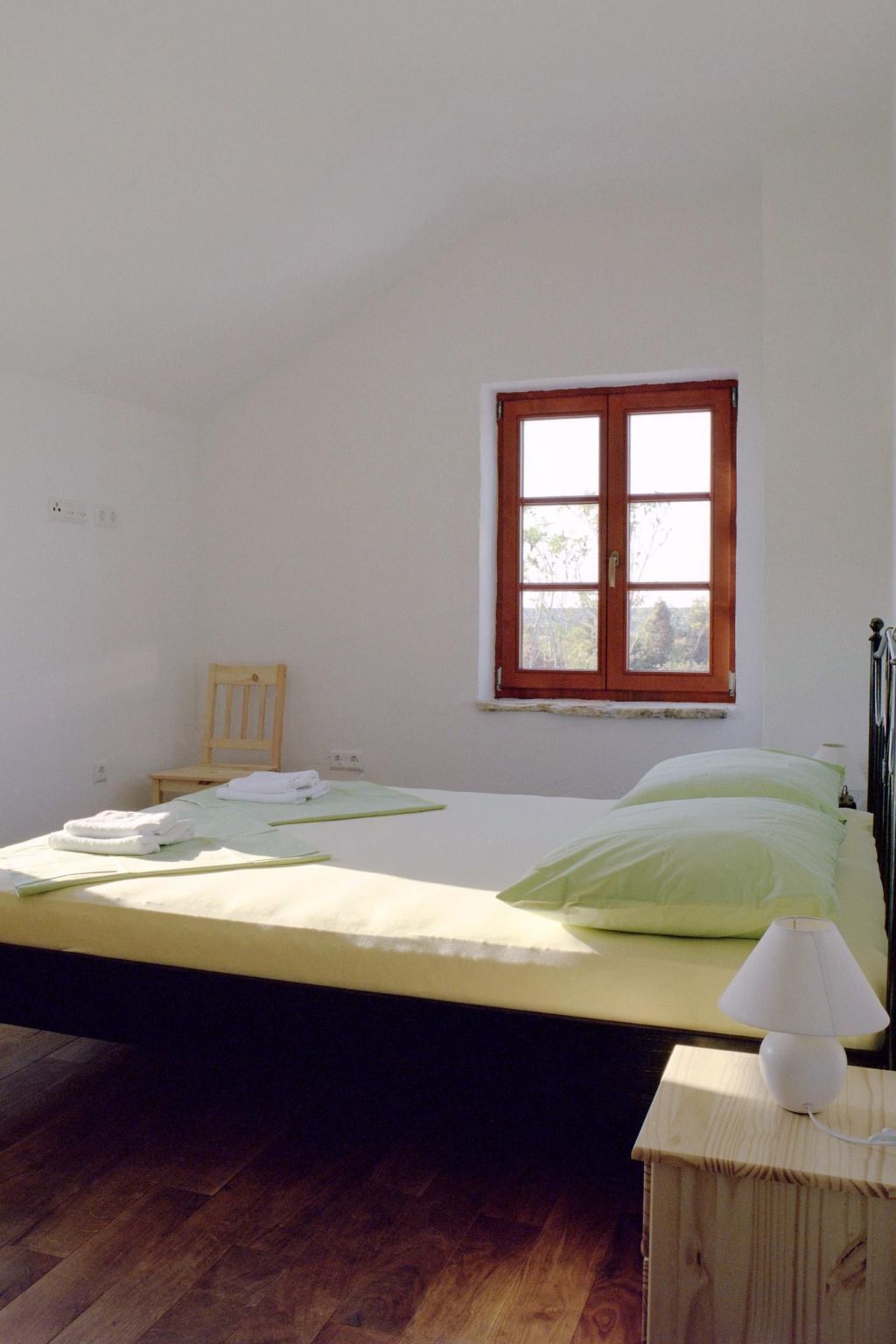 A bedroom with a single bed, a wooden nightstand, a lamp, and a window with a view of trees.