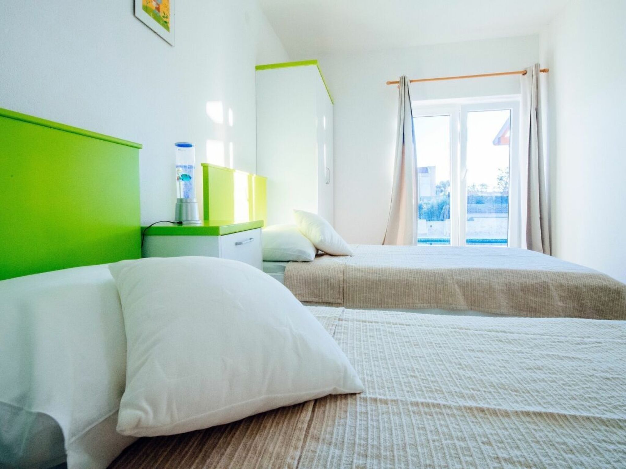 Une chambre d’hôtel avec deux lits, une tête de lit verte, une table de chevet avec une bouteille d’eau et une fenêtre avec des rideaux.