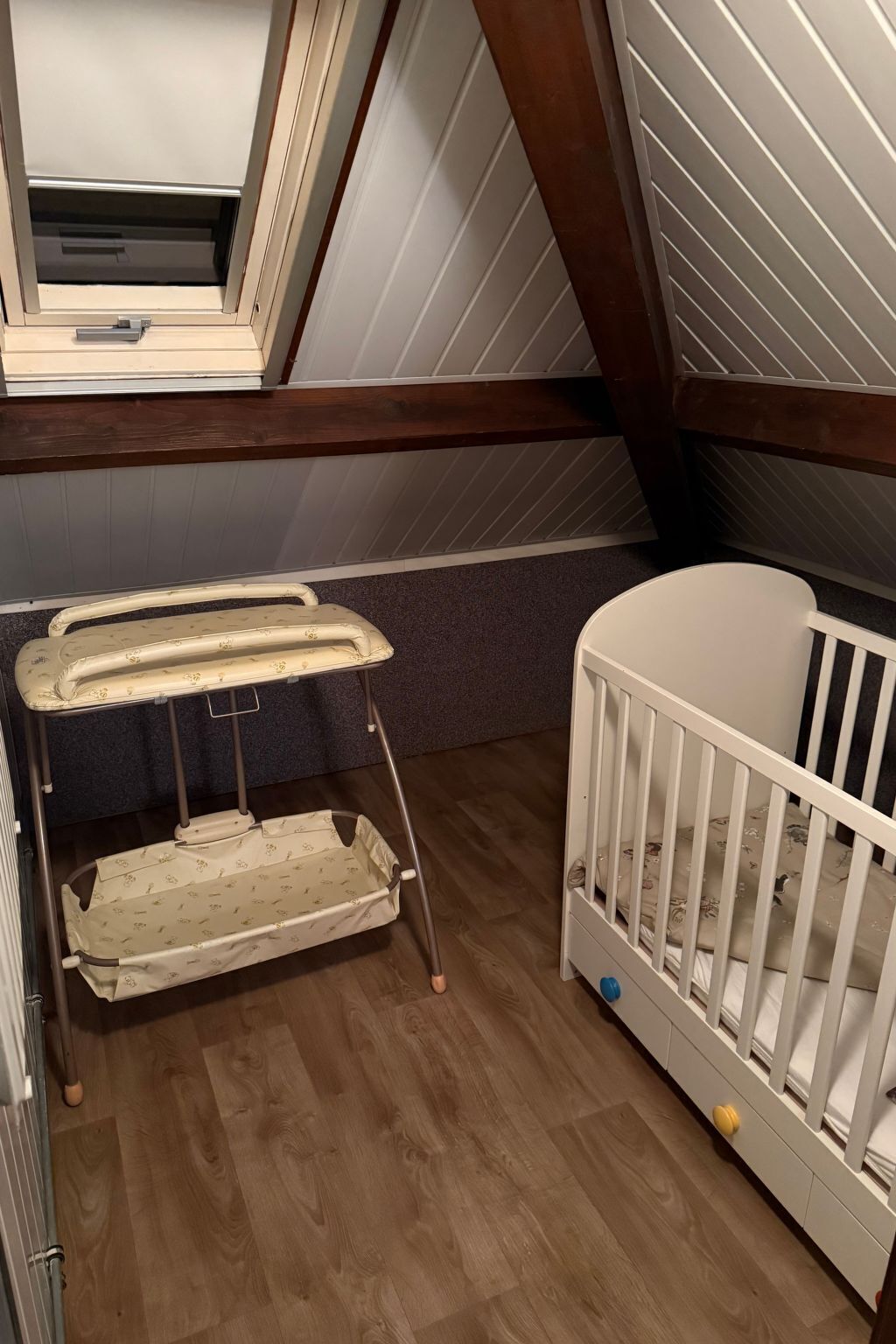 A room with a crib, a changing table, and a window with a view of the roof.