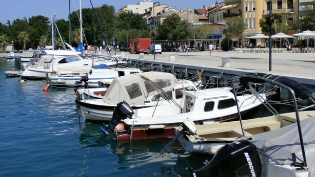 Ein Yachthafen mit verschiedenen Schiffen an den Liegen, einer Uferpromenade und Gebäuden im Hintergrund.