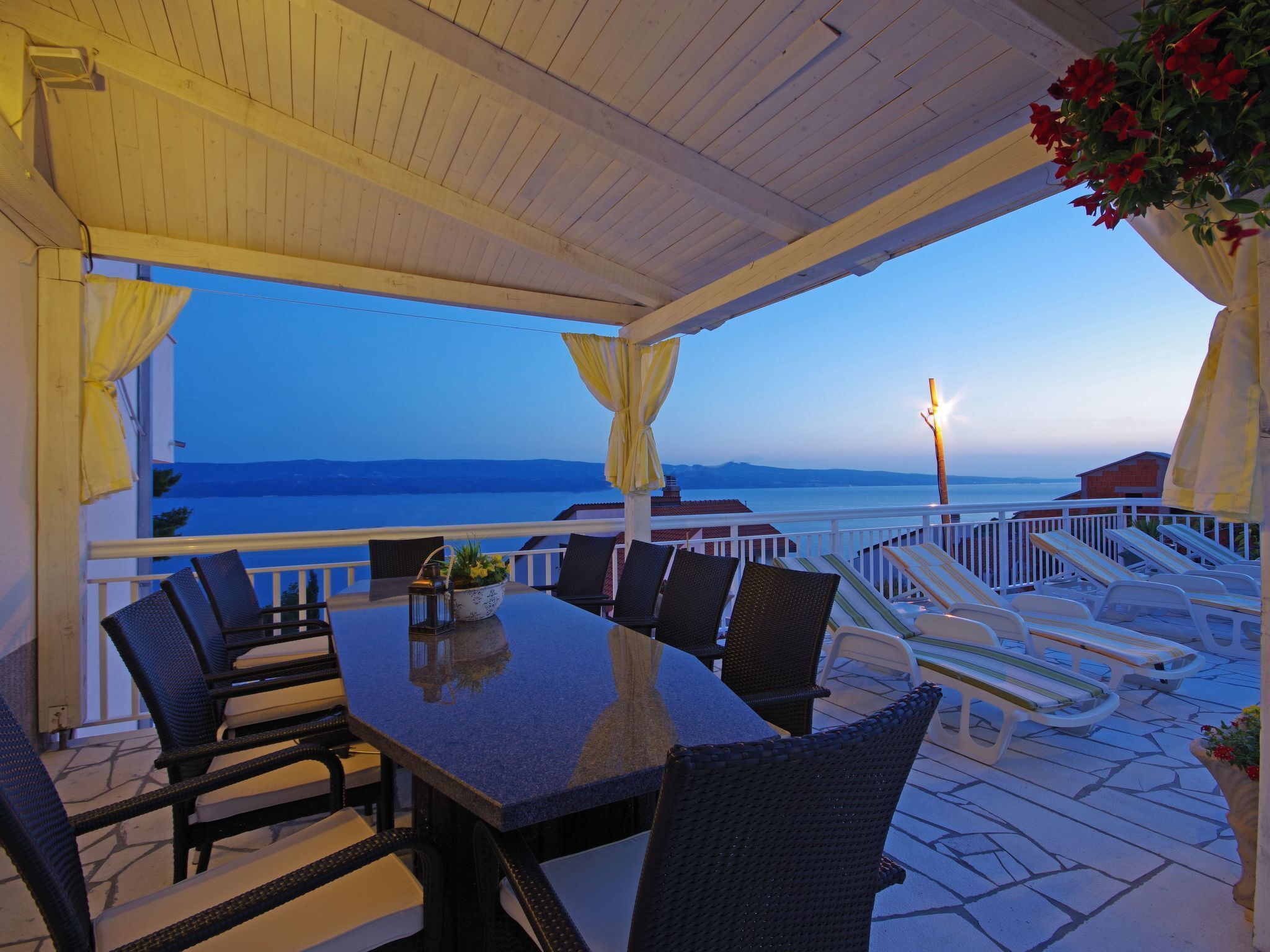 A covered outdoor seating area with a table and chairs, overlooking a body of water and distant mountains.
