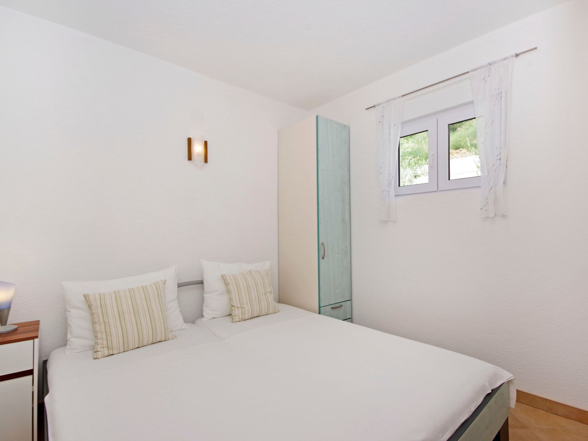 Ein Schlafzimmer mit einem Bett, einem Nachttisch, einem Fenster mit Vorhängen und einer Wandleuchte.
