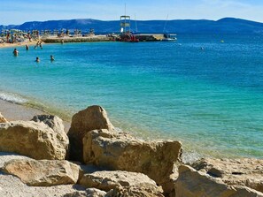 Ein Strand mit klarem türkisblauem Wasser, einem felsigen Vordergrund und einem Steg mit Menschen und Booten.