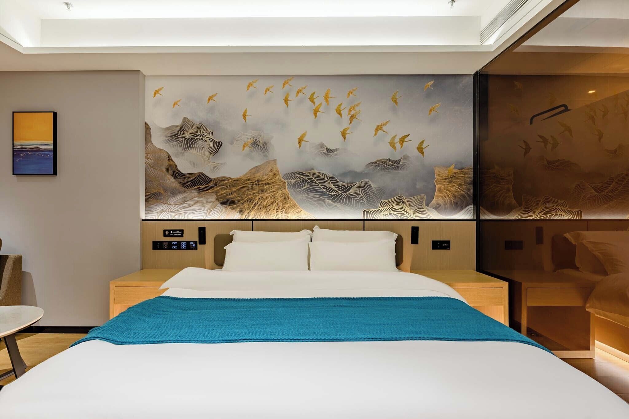 Une chambre d’hôtel moderne avec un grand lit, une table de chevet, une chaise et une œuvre d’art fixée au mur représentant des oiseaux et des montagnes.
