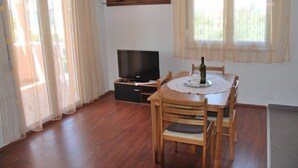 Habitació amb taula de menjador de fusta, cadires, un televisor sobre un suport i una ampolla sobre la taula.